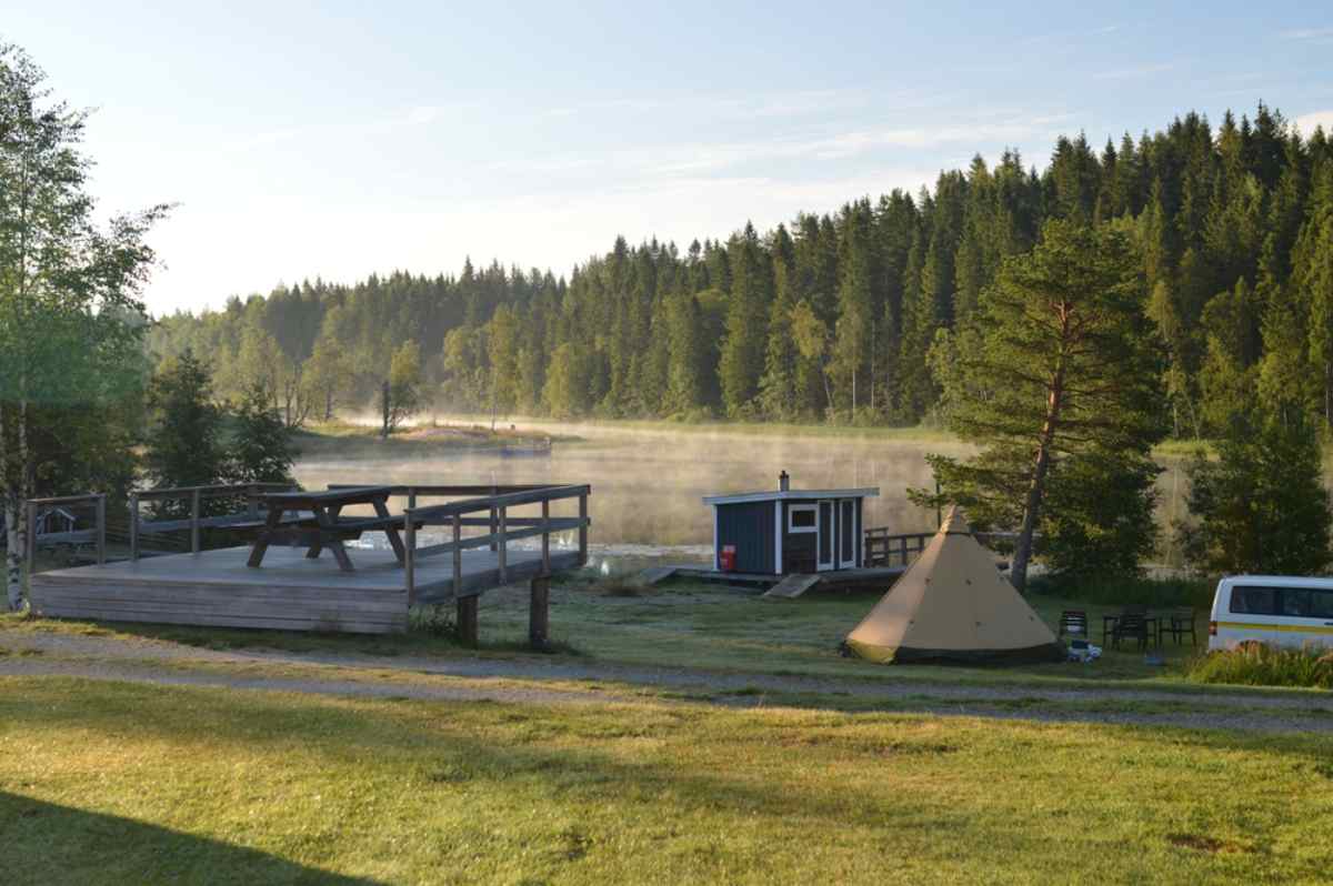 General view of a Swedish campsite for campers near what looks like a lake surrounded by tall trees.